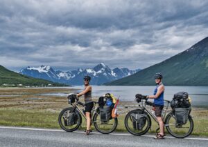 A man and woman posing with their loaded touring bicycles in front of a lake and mountains.