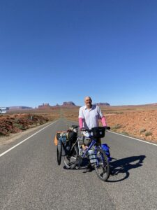 A man with a fully loaded touring bicycle standing on a deserted highway in the desert.