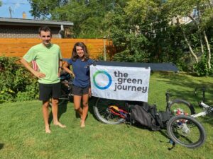 2 people holding a sign that says The Green Journey standing in front of a touring bicycle