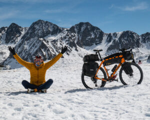 man sitting in the snow with outstretched arms next to a loaded touring bicycle