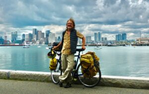 A man posing with his fully loaded touring bike on a path with a lake and city skyline in the background