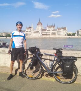 Man standing next to his touring bicycle in front of a river with a castle behind it