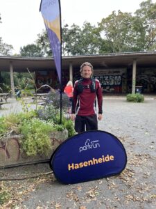 Man dressed in bicycling gear standing in front of a Parkrun event sign