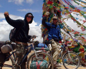 2 men with fists raised on their bicycles in front of ceremonial flags